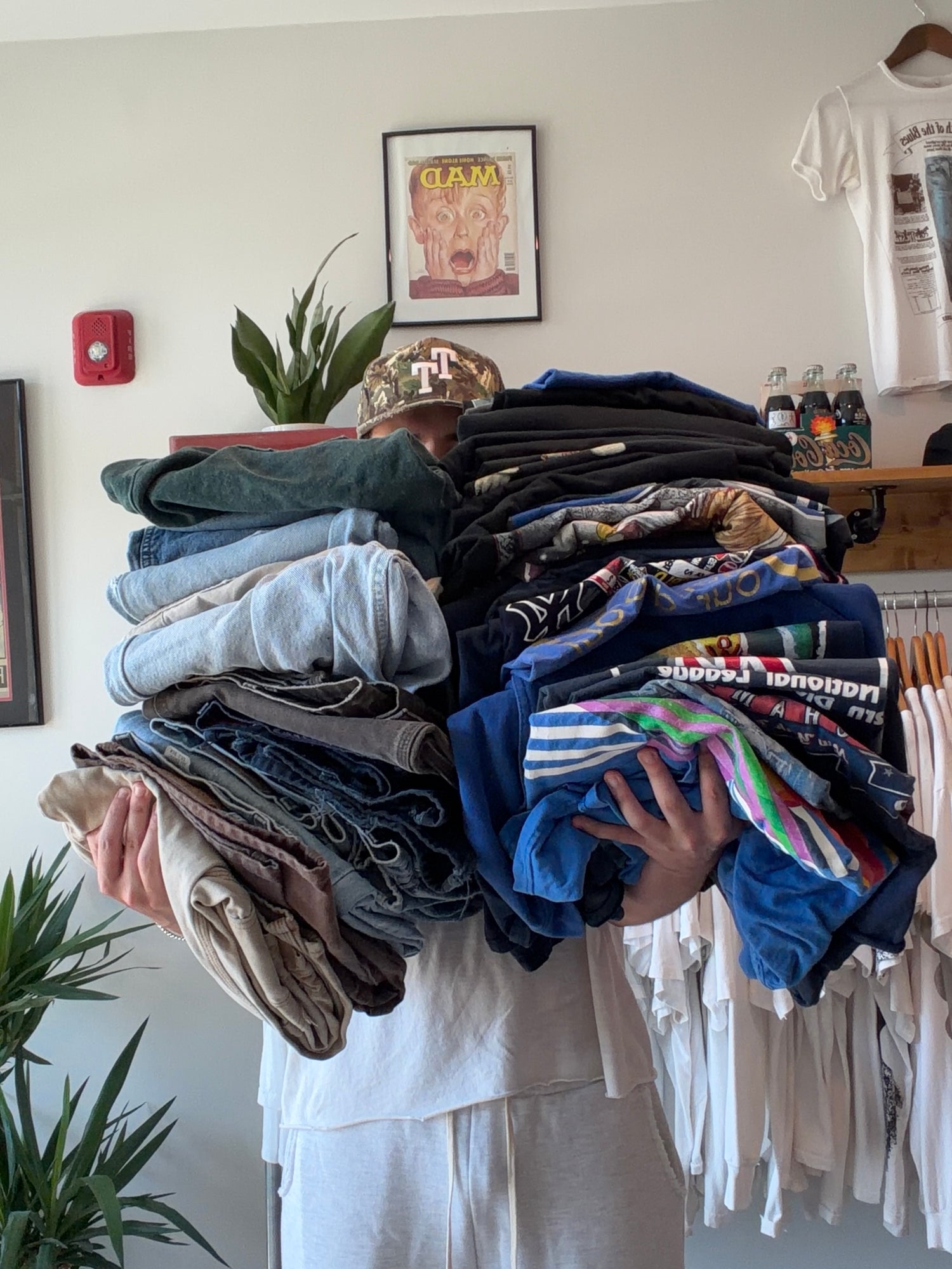 A person in a white outfit and camouflage cap carries a large stack of folded clothing, including jeans, t-shirts, and patterned tops, in an indoor room with shelves, plants, and hanging garments visible.